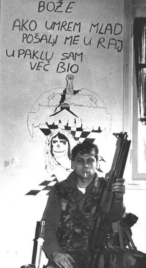 Croatian soldier (1992)in front of a wall that says , “God if I die young send me to Heaven as Hell I have already been”
