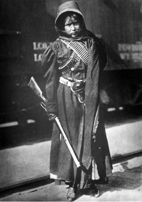 Mexican female soldier (Soldadera) stares down the camera while holding her riffle. Behind is the train before shipment to war, 1914.