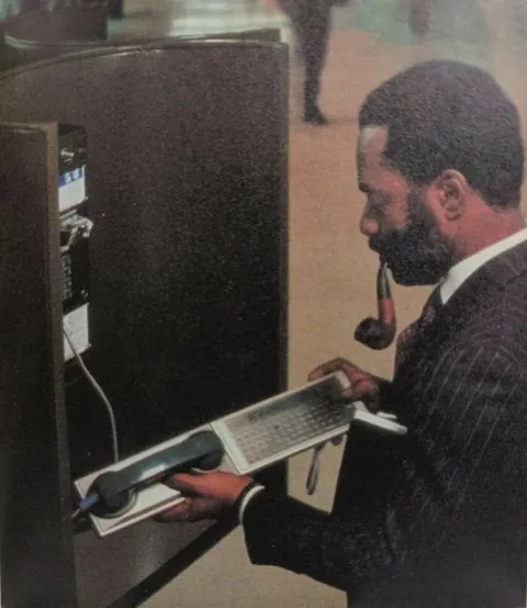 Man Checks His E-Mail Over A Public Pay Telephone Using A Panasonic Rl-P4001 Acoustic Coupler Dial-Up Modem Attached To A Panasonic Rl-H1400 Hhc (Hand-Held Computer) In The Early 1980s