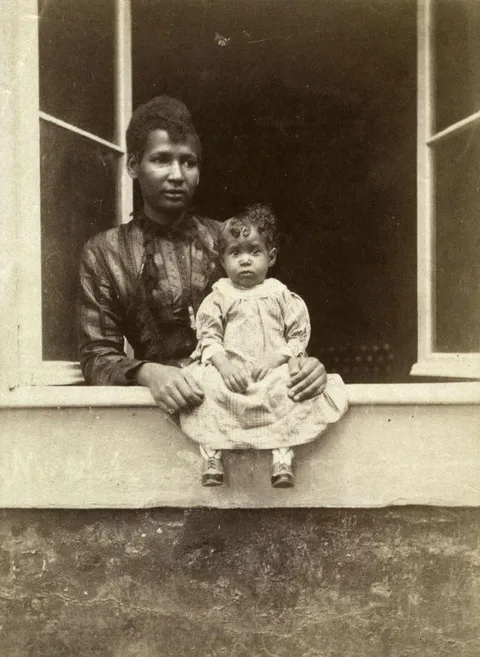 Young mother poses her baby thru the window of her home. Circa 1880s.