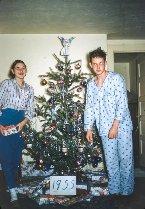 Sister smiles (almost laughs) as she poses with her christmas tree, her brother tries to stand half dead-sleep, December of 1955.