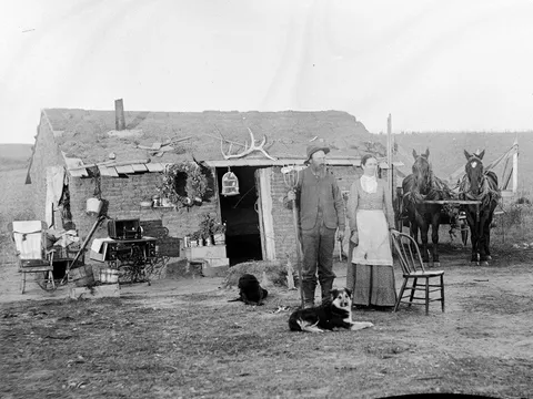 A married couple on the Nebraska prairie, 1800s. Though ramshackle and leaning a little, their house has homey touches, including a decorative wreath and some pet songbirds in a cage.