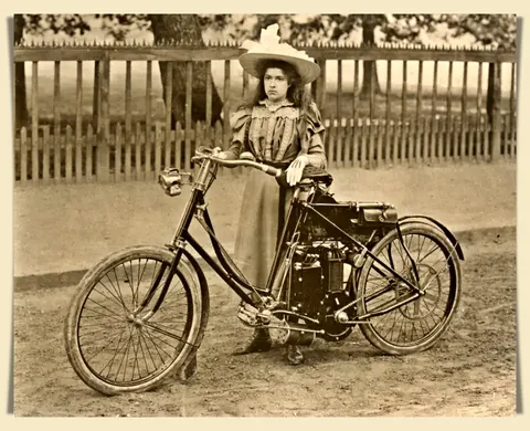 Young lady posing with a very early Lawson motorcycle model, with a top speed of 19 km/h. circa 1897.