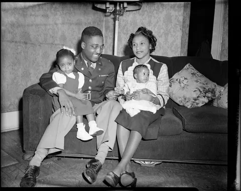 Father proudly sits in full uniform with his family, trying to get the attention of his smallest child, safety film, 1940s.