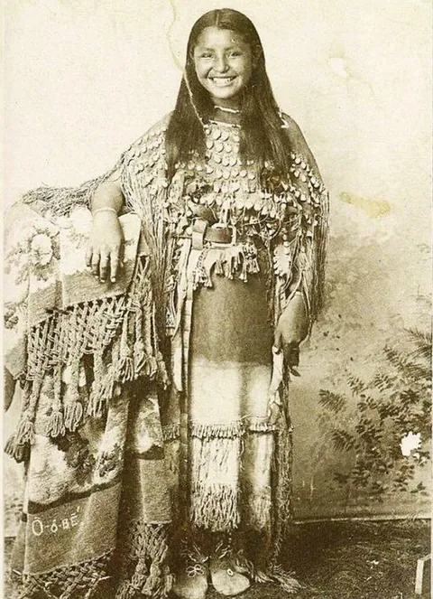 Portrait of O-o-be, a Kiowa girl wearing a dress decorated with elk teeth. Her name is embroidered in the textile next to her. Photographed in 1895. Smithsonian Museum.