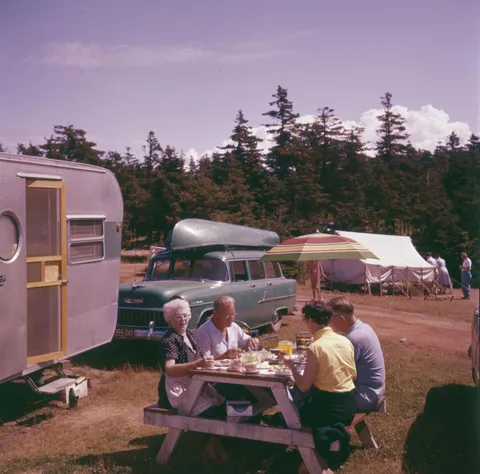 "Two men and two women eating at a picnic table in front of a car with a canoe and a trailer home, Prince Edward Island", 1956. Photo by Chris Lund.