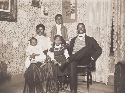African american family poses in 1904. Father tries to look dignified while mother gives a bright smile.