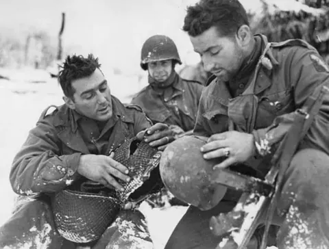 During the Battle of the Bulge, PFC Andy Masiero &amp; Staff Sgt Urban Minicozzi of the 75th Infantry Division examine the headgear of PFC Masiero in the Ardennes. The young private’s helmet was pierced by an enemy’s bullet which drew blood from his temple, but left him otherwise unscathed. (1944)