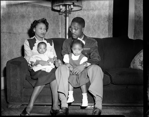 Father proudly sits in full uniform with his family, trying to get the attention of his smallest child, safety film, 1940s.